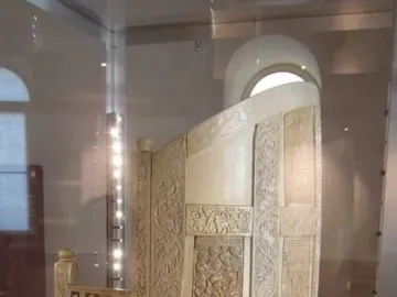 Throne of Maximian with a high arched back and ornate relief carvings inside a glass case.