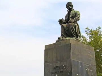 Bronze monument of Vuk Karadžić seated on a tall stone pedestal, holding a book, with trees and a cloudy sky behind it.