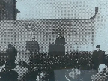 The Angel of Peace monument stands on a pedestal behind a man speaking at a podium to a crowd.