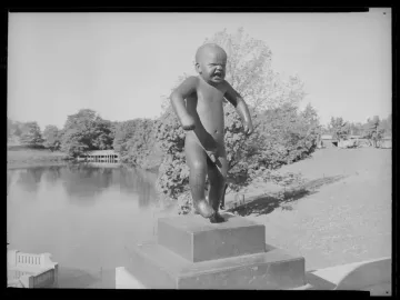 The Angry Boy sculpture stands on a pedestal outdoors beside water and trees.