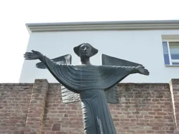 Angel of Peace statue with outstretched arms on a reddish stone pedestal, set against a brick wall and modern building.