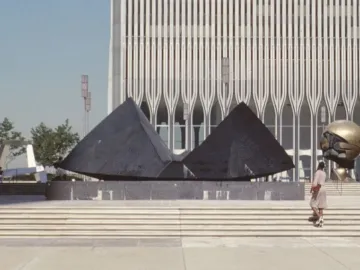 Cloud Fortress sculpture stands in a plaza before a tall ridged tower, with a spherical sculpture partly visible at right.