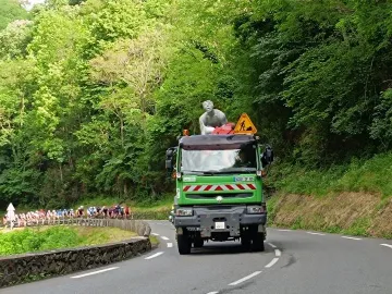 The Géant du Tourmalet statue mounted on a green utility truck on a winding mountain road, with cyclists in the distance.