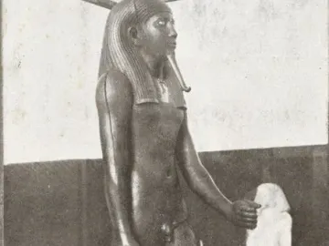 Statue of King Hor standing in a museum display, wearing a striped headdress and false beard and holding a staff.