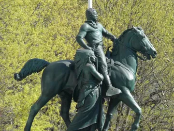 Theodore Roosevelt Equestrian Statue with a horse, a walking figure in a feathered headdress, and leafy green trees behind it.