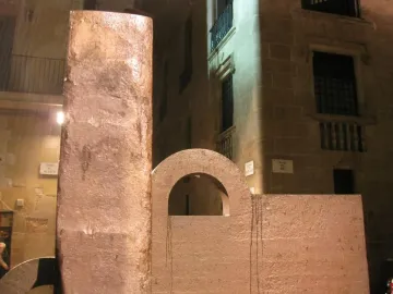 Topos V sculpture by Chillida with a tall pillar and an arched block against historic stone buildings.