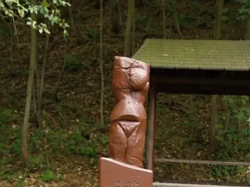 Venus of Petrkovice sculpture on an inscribed plinth and stone-filled gabion base in a wooded setting.