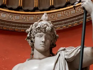Marble statue of Antinous Braschi with a leaf and grape wreath, bare chest, draped cloth, and a staff against a red background.