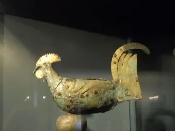 Gallo di Ramperto rooster sculpture with a weathered greenish-brown patina on a tall stand.