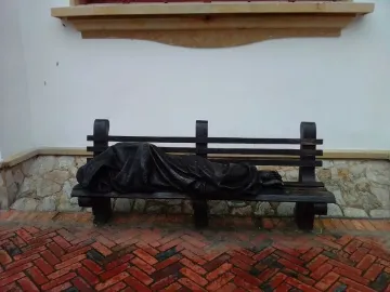 Jesus the Homeless sculpture of a draped figure lying on a park bench against a white wall.