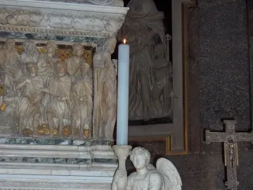 Kneeling white marble angel holding a tall candlestick with a lit candle, set before an ornate relief.