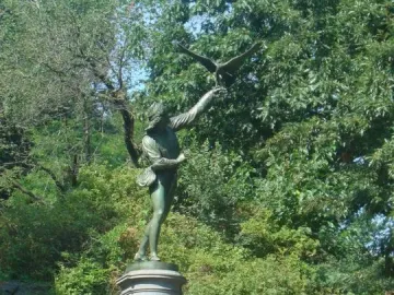 The Falconer bronze statue of a man releasing a bird of prey on a stone pedestal among green foliage.
