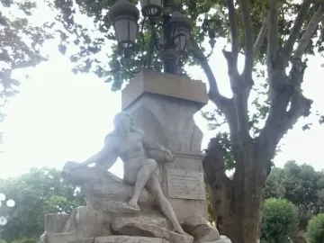 Ain El Fouara Fountain with a reclining nude female statue on a rocky base and a lamppost above it, framed by a leafy tree.