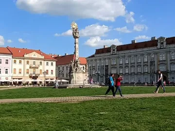 The Holy Trinity Monument stands in a sunny public square beside historic buildings and a grassy area with people walking nearby.