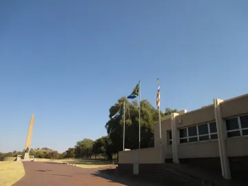 The National Women\'s Monument stands on a raised base in a grassy landscape with a paved path leading toward it and a modern building nearby.