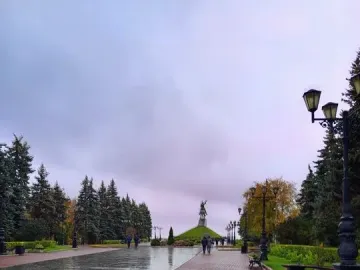 A paved park path leads toward the Salavat Yulaev Monument on a grassy mound, lined with trees and street lamps under an overcast sky.