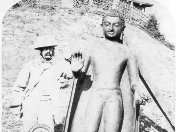 A man in colonial-era clothing and a pith helmet stands beside the Sultanganj Buddha, with two smaller carved stelae at the base.
