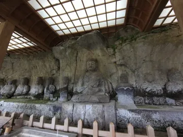 Usuki Stone Buddhas carved into a rock cliff beneath a protective wooden shelter with a fenced viewing area.