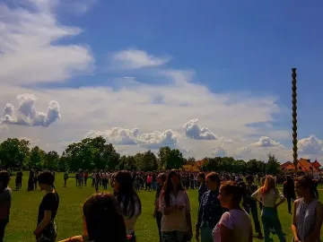 The Endless Column rises beside a green field under a blue sky with clouds, with many people gathered below.