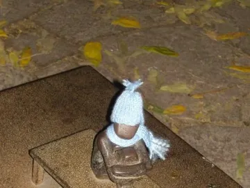 Järnpojke, a small dark sculpture of a boy wearing a light blue knitted hat and scarf on a tiny bench, with coins nearby.