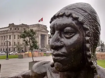 Close-up of La Yunta bronze sculpture with a classical building and Peruvian flag in the background.