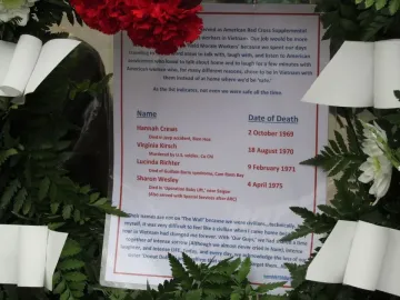 A laminated sign with names and text, framed by green leaves, white ribbons, and red flowers at the Vietnam Women\'s Memorial.