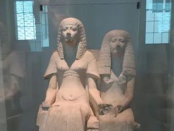 Horemheb and Amenia seated together on a shared throne, holding hands in a stone sculpture.