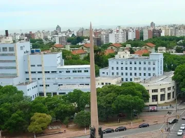 The Obelisk of Montevideo rises from a hexagonal water basin in the center of a busy traffic roundabout, with city buildings and trees around it.