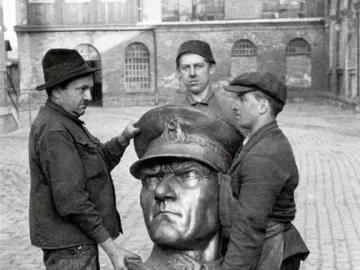 Three workers carrying a large bronze head sculpture of Atatürk in a courtyard.