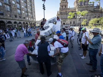 People gather in a sunlit public square around a wrapped structure being loaded onto a flatbed, with a cathedral tower in the background.