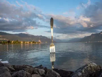 The Fork of Vevey sculpture stands in the lake with mountains and a cloudy sky in the background.
