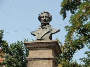 Dark bust of Pushkin on a stone pedestal outdoors, framed by trees against a blue sky.