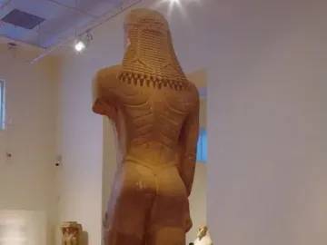 Rear view of the Sounion Kouros, a colossal ancient Greek stone statue of a nude male figure in a museum gallery.