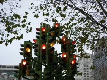 Traffic Light Tree sculpture made of green poles and many illuminated traffic lights in Canary Wharf, with buildings and vehicles behind it.