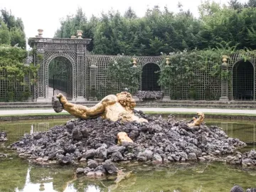 Enceladus Fountain showing a golden muscular figure rising from dark rocks in a greenish pool, with an arched trellis and greenery behind.