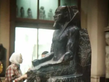 Khafre Enthroned seated dark pharaonic statue with a false beard in a museum, shown beside a person for scale.