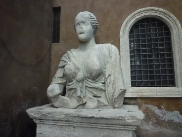 Madama Lucrezia, a weathered white marble statue of a woman in classical drapery on a stone pedestal beside a building wall and arched window.