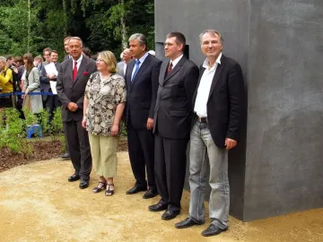 Five people stand beside the Memorial to Homosexuals Persecuted Under Nazism on a gravel path with trees and a crowd in the background.