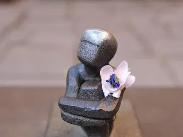 Järnpojke sculpture with a shiny head, a small flower in its chest, and coins scattered in front