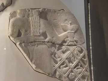 Fragmented relief from the Stele of the Vultures showing a large figure holding a staff with a bird emblem above smaller figures trapped in a net, with cuneiform text above.