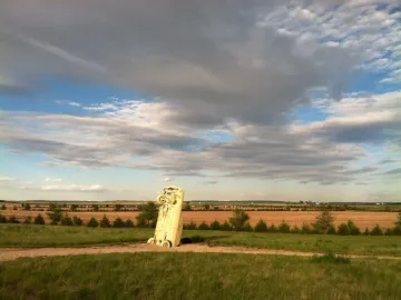 Carhenge sculpture with a pale vintage car standing vertically in a grassy field under a cloudy sky.