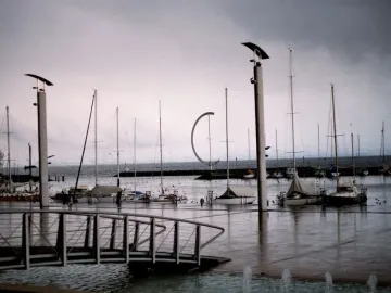 Éole, a dark C-shaped metal sculpture, stands on a breakwater near sailboats in the marina.