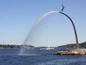 God our Father on the Rainbow sculpture rising from the water, with a sailboat nearby and islands in the distance.