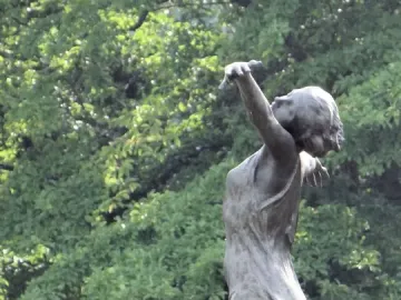 The Dancing Girl sculpture in dark metal, seen from a low angle among blurred green foliage.