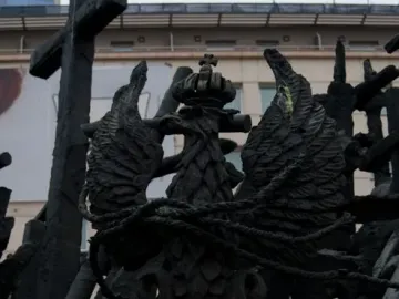 Dark metal memorial sculpture with a crowned eagle emblem, a large cross, and a ribbon tied at the base.