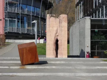 Pi-Chacán stone sculpture with a deep vertical crevice, standing beside a rusted rectangular block on a paved area.