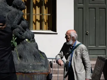 Pillar of Shame sculpture with white roses at its base, flanked by two men observing it