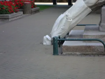 Rolleston Statue toppled over on a paved walkway beside a stone base, with red flowers and a green railing in the background.