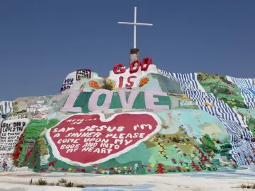 Salvation Mountain covered in colorful religious murals, with a white cross on top and large painted words including LOVE and GOD IS.