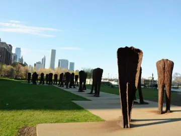Agora sculpture with dozens of rusted, headless human-like figures spread across a grassy area and walkway in front of the Chicago skyline.